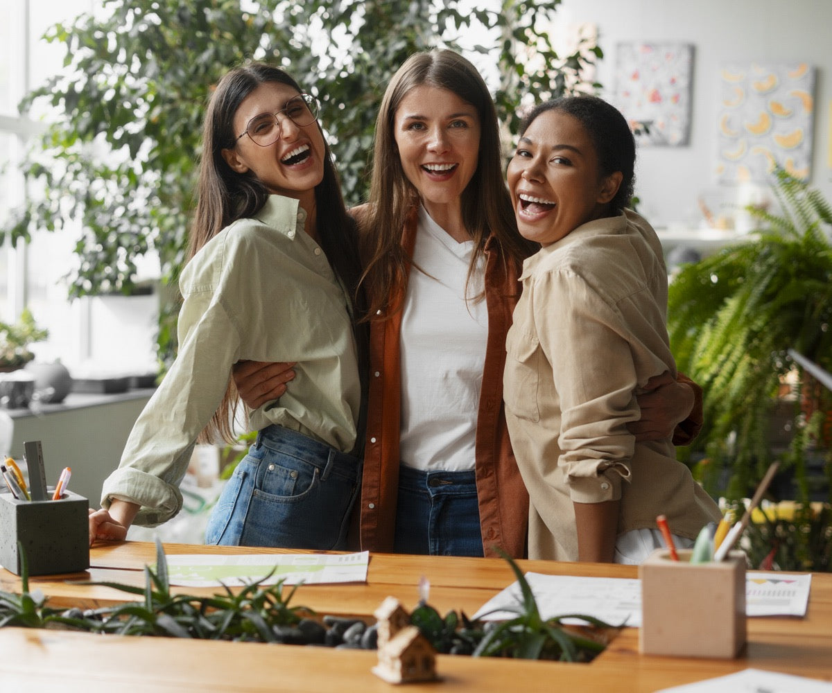 Trois femmes souriantes partagent un moment de rire à un bureau, illustrant une ambiance de travail agréable.